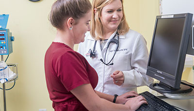 Nursing student typing at a computer while a professor looks on.