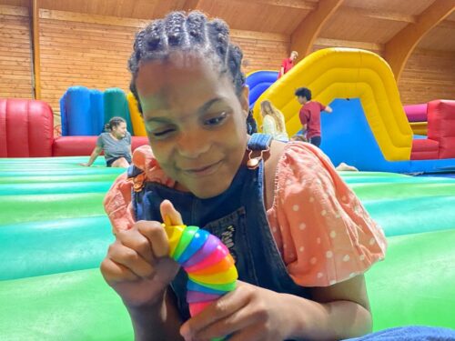 On a big inflatable play area, a girl with braided hair, wearing an orange top and dungarees plays with a colourful toy.