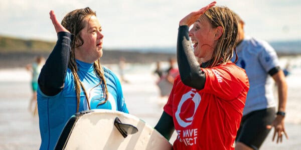 Two women in wetsuits hold surfboards in one hand and high five with the other.