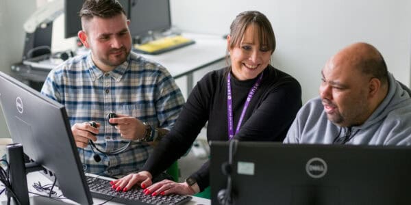 Two men sit either side of a woman at a computer in an office, all laughing and chatting together.