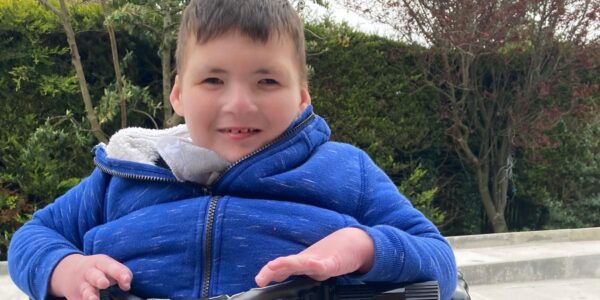 A boy wearing a blue coat uses a mobility device while smiling