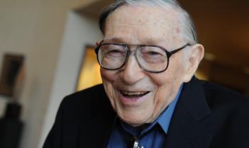 Older man smiling with gray hair and dark glasses wearing blue shirt