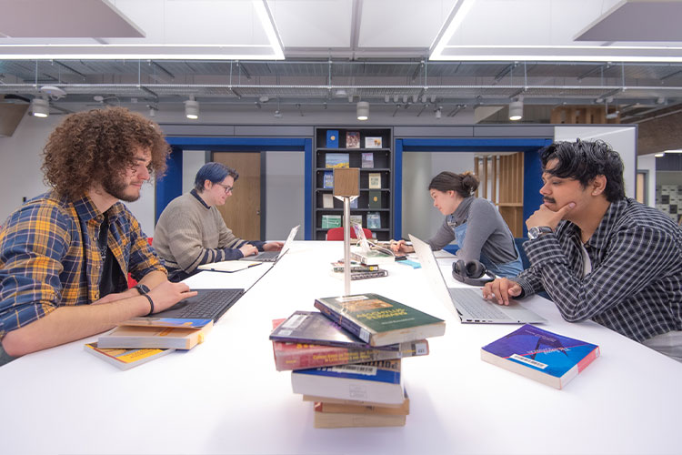 Students sitting at a library study desk with books and laptops