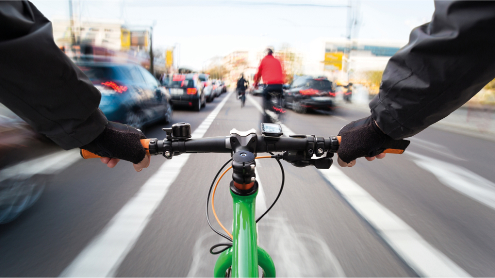 Blick eines Radfahrenden mit einem grünen Fahrrad auf einem Radfahrstreifen. Vor dem Radfahrenden sind weitere Radfahrende zu sehen, rechts und links Autos.