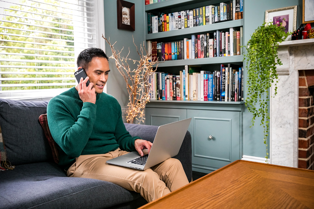 A man is sat on a sofa, speaking on a mobile phone while looking at his laptop