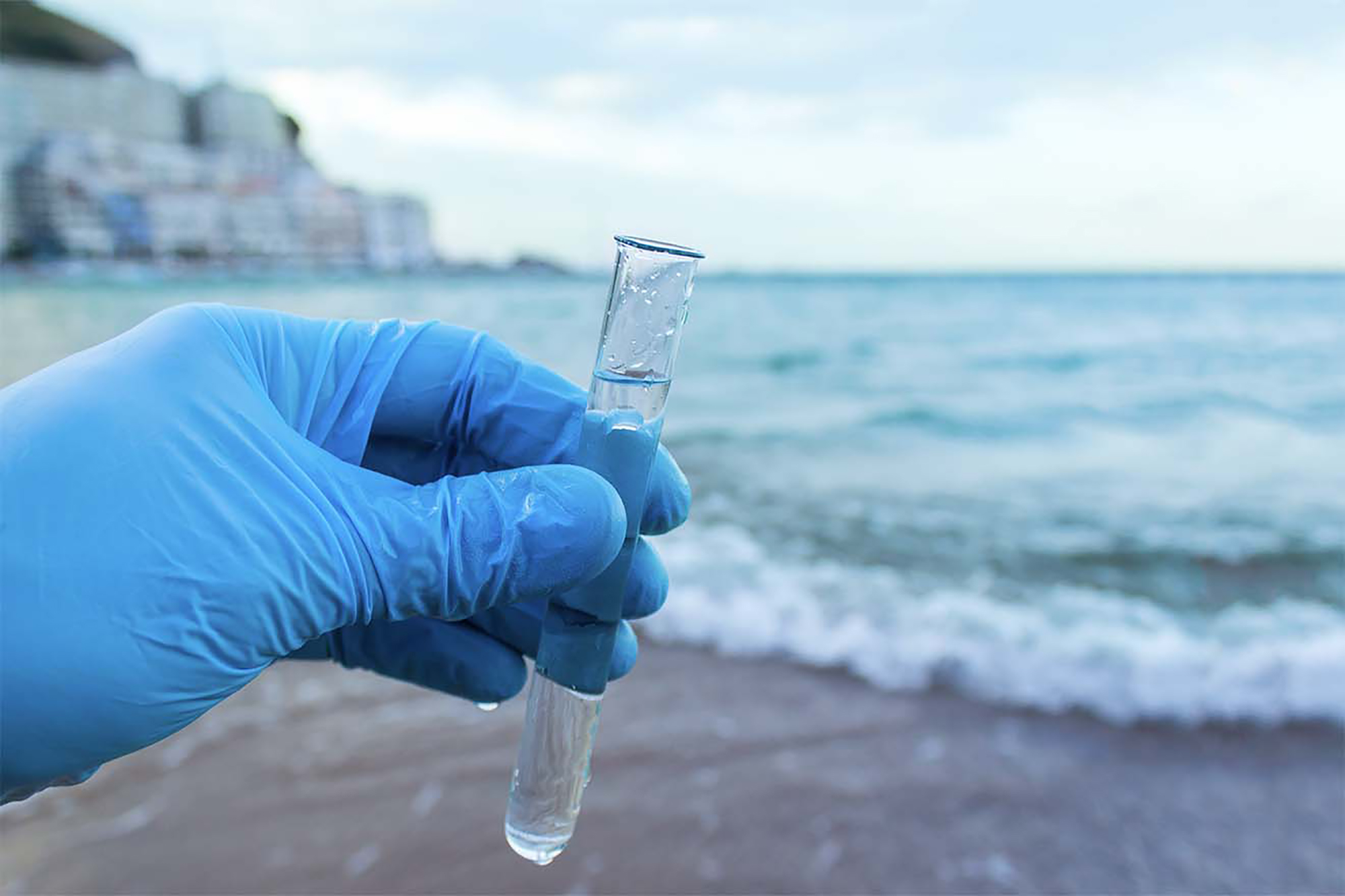 A hand wearing a glove holds a test tube on a beach.