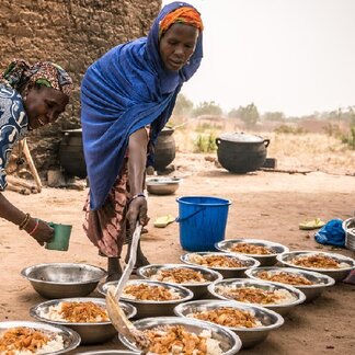 Mothers are dishing out the freshly prepared school lunch onto large plates. The meal is a large portion of rice with beans and vegetable stew on this day.