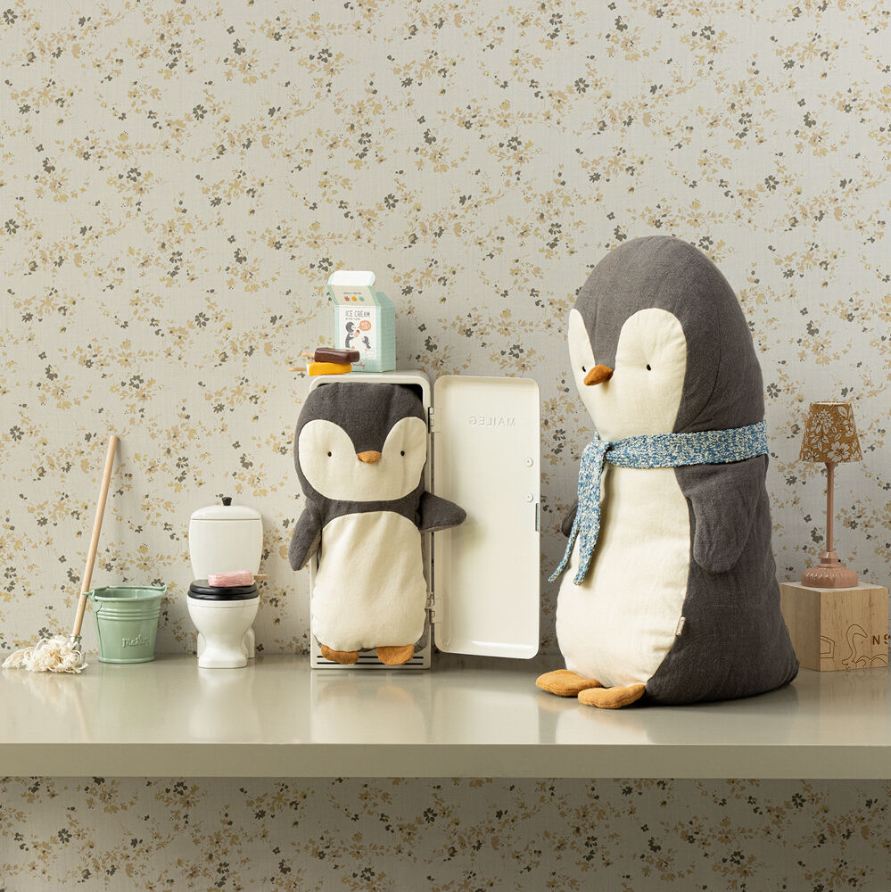 Ein graues und weißes Kuscheltier in Form eines Pinguins mit einem blauen Schal sitzt auf einem Tisch vor einer gemusterten Tapete. Links daneben steht ein kleinerer Pinguin.