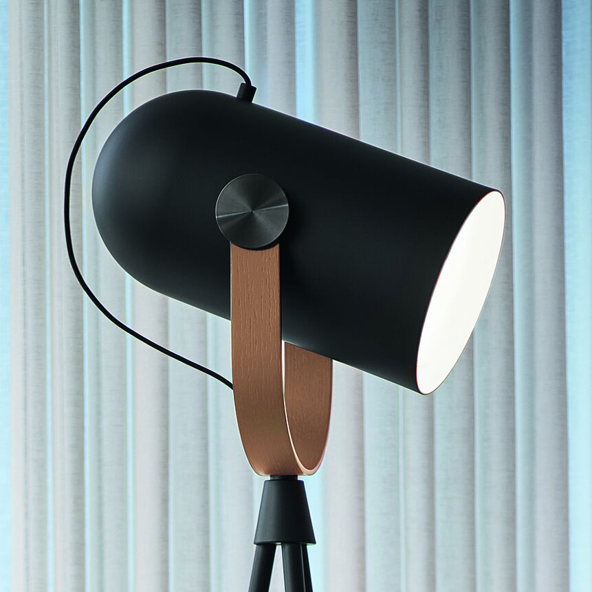 Die ZEIT Sonderedition Stehleuchte 'Carronade II' zeigt eine moderne Stehlampe im skandinavischen Design. Sie hat ein schwarzes Dreibein-Gestell mit einem verstellbaren Lampenkopf, der von Holzdetails akzentuiert wird. Die Lampe steht vor einem hellen Vorhang in einem modernen Wohnbereich.