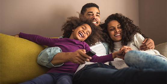 A family of three smiling and cozying up on a couch together while watching TV.