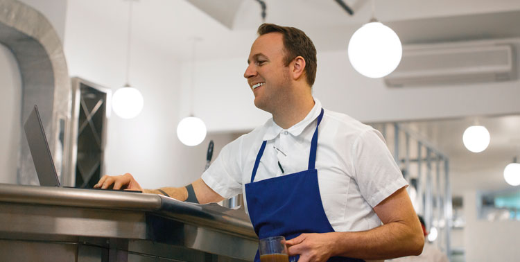 A man with a blue apron smiles while holding his coffee and working on a laptop computer.