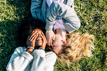 Two people laying on the grass. One is whispering in the other one’s ear.