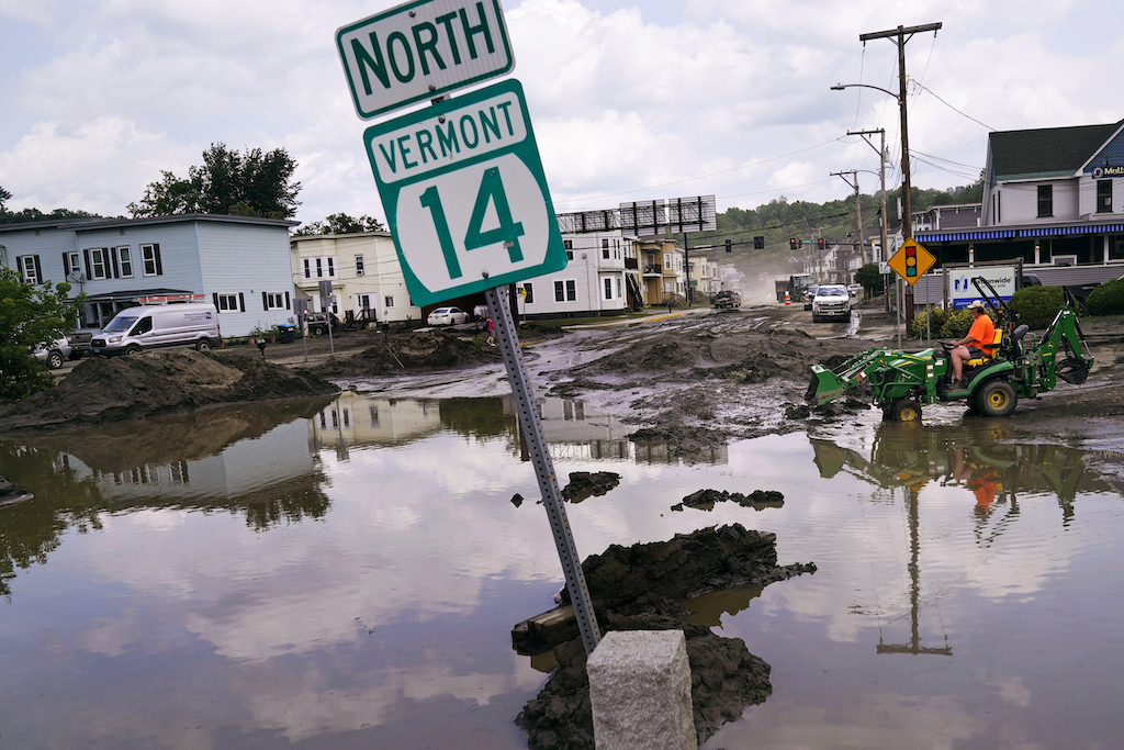 A small tractor clears water from a business as floodwaters block a street in Barre, Vt., in July 2023. Heavy flooding last year devastated many Vermont communities. Now, state lawmakers are backing a “climate Superfund” bill that would force fossil fuel companies to pay for climate change-caused damages based on their emissions. (Photo by Charles Krupa/The Associated Press)