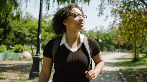 nicole, an amazon future engineer scholarship recipient, walks down a campus road while wearing a backpack. she's wearing glasses and is looking off camera to her left