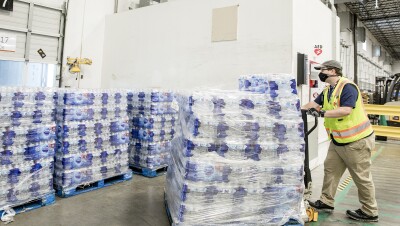 A man wearing a yellow safety vest and face mask maneuvers a pallet of water bottles in a warehouse setting. 