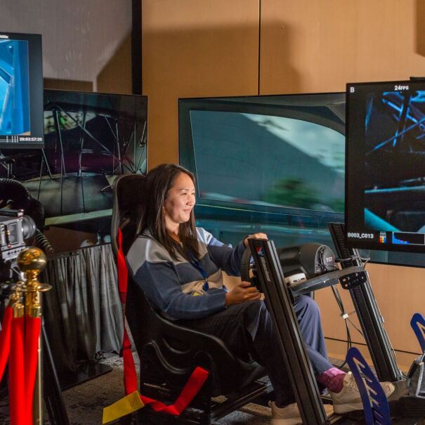 author connie chen sitting in a car shell pretending to drive a car while her face is projected onto screens that show her racing around a racetrack