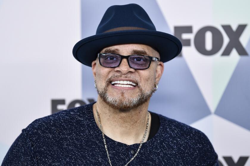 Sinbad in a black wide-brimmed hat, black shirt, necklace with a cross pendant and sunglasses smiling