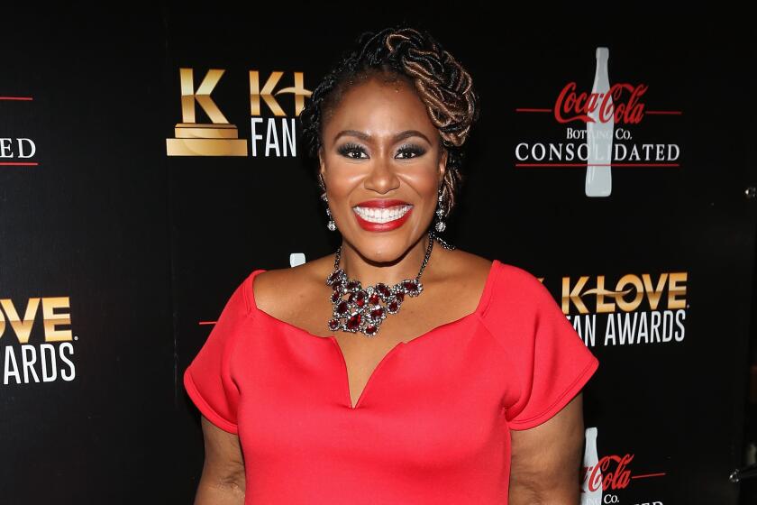 A Black woman in a red gown with hair in a braided style and a necklace smiling against a black background