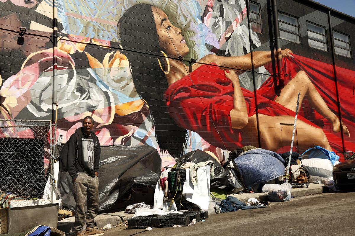 A man stands in front of a row of tents and makeshift enclosures and a large mural on Wilde Street in Skid Row