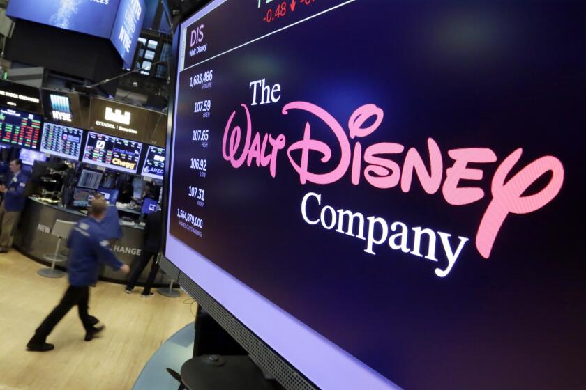 A pink-and-white logo for the Walt Disney Company appears on a screen in an office with other screens in the background.