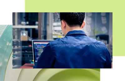 Rear view of a man in a blue work uniform using a laptop in an industrial setting.