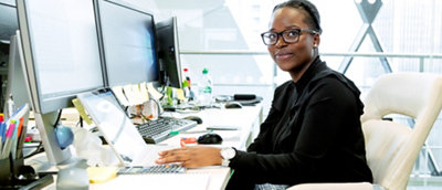 A woman in glasses and a black shirt sits at a desk with multiple computer monitors in a well-lit office.