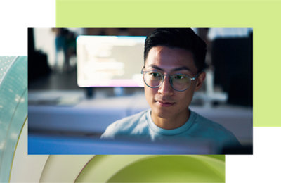 Young asian man wearing glasses, looking at the camera, with a computer screen glowing in the background.