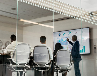 Four professionals in a meeting room, one pointing at digital data displayed on a large screen, viewed through a glass wall.