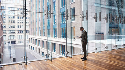 高いビルの中に、大きなガラス壁の前に立ってスマートフォンを使っている人。