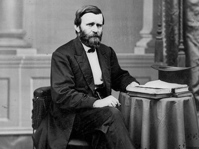 Full-length portrait of Ulysses S. Grant seated at table with books and top hat, facing right, ca. 1869-1877.