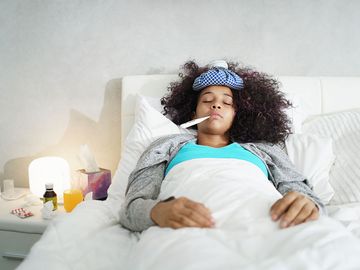 Ill young black (african american) woman with cold, lying in bed and holding a thermometer in her mouth.