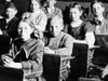 Vintage image of students at desks in a classroom with a teacher standing in the background. (education, learning)