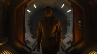 a person in an orange flight suit walks down a hexagonal hallway in a dark spacecraft
