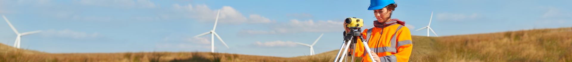 a female wind farm engineer at work wearing a high visibility jacket and a blue hard hat. There are wind turbines across the landscape in the background.