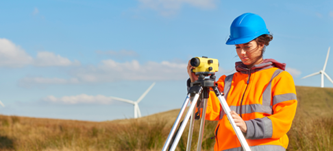 a female wind farm engineer at work wearing a high visibility jacket and a blue hard hat. There are wind turbines across the landscape in the background.