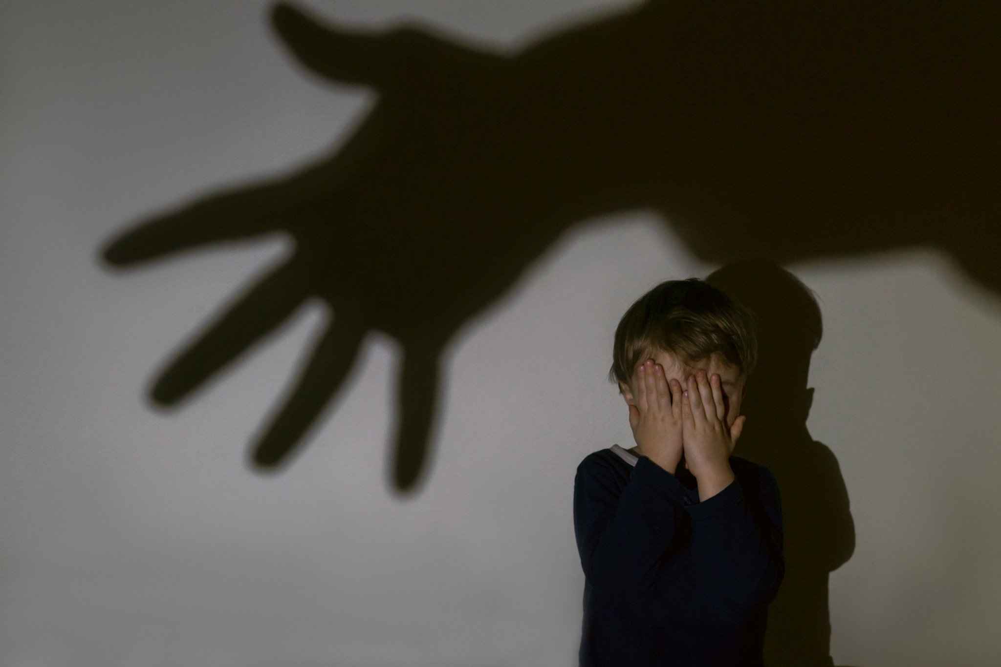 A little boy and scary shadow of hand on a light background for a fear or kidnapping concept. Boy closed eyes with hands.