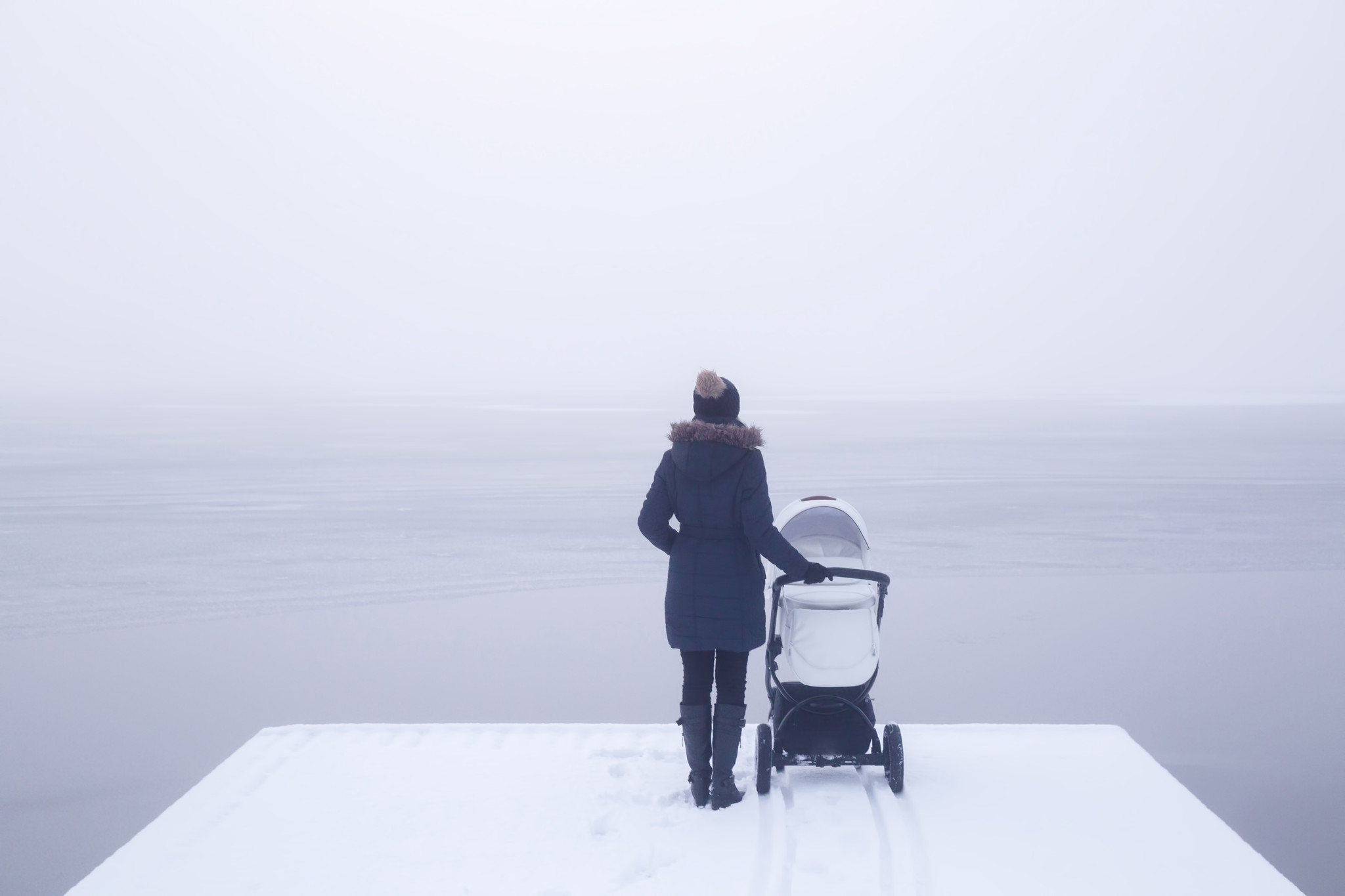 Young mother standing with baby stroller on snow covered footbridge and staring at lake. Mist over frozen water. Foggy air. Winter day. Back view. Empty place for sentimental text, quote or sayings.