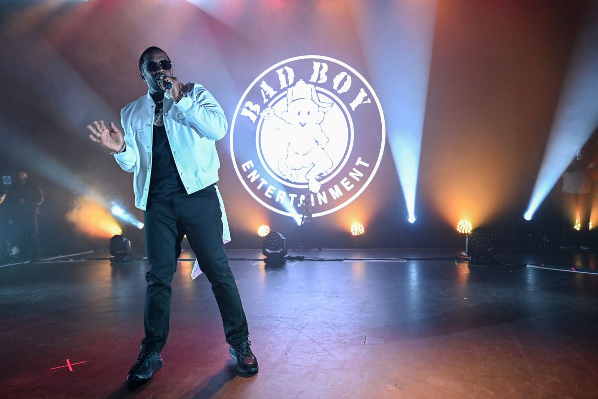 Diddy performs onstage wearing black clothes, a white jacket, and sunglasses. A Bad Boy Entertainment logo is projected on the wall behind him.