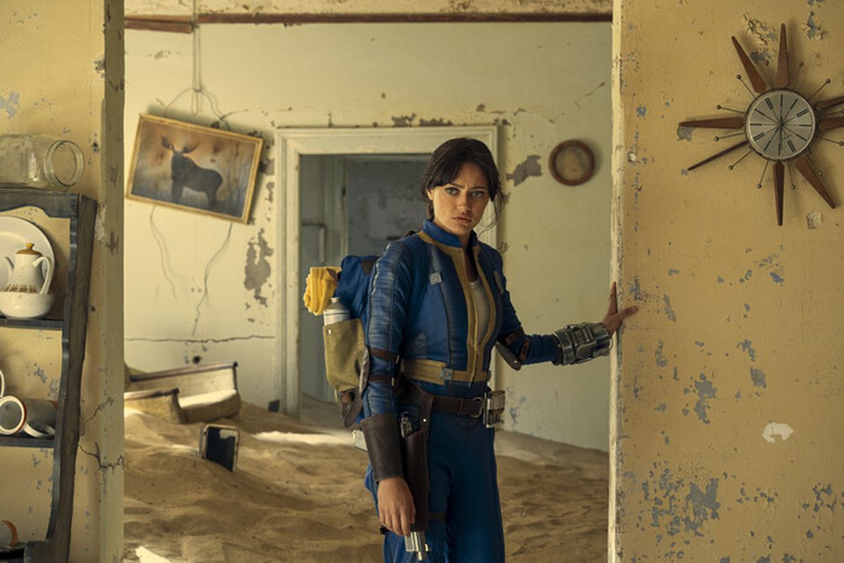 A young woman wearing a backpack stands in the doorway of a derelict room that appears to be abandoned. Wall decorations hang askew and the wall paint is chipped and damaged. The ground is covered in sand.