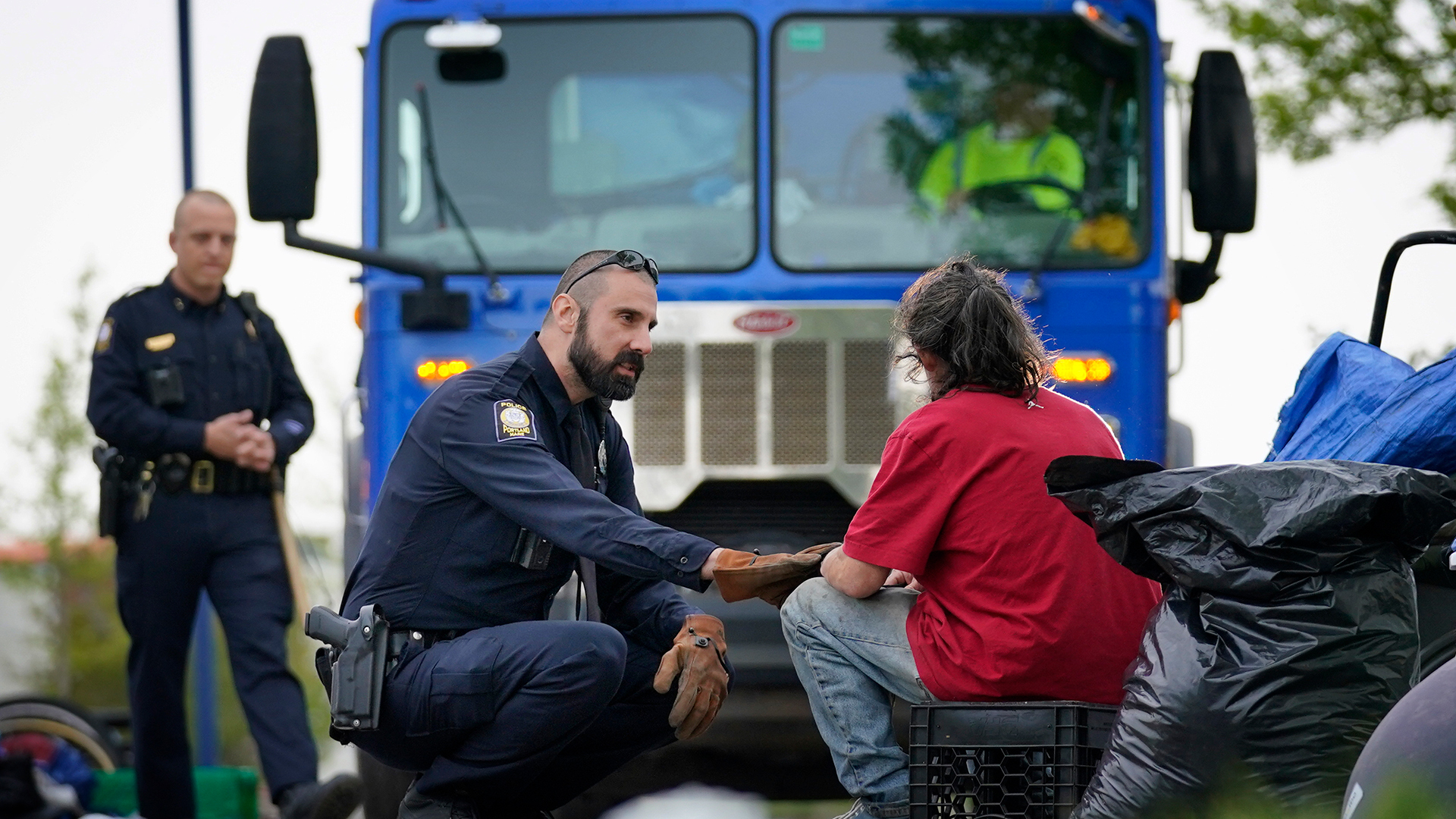 A police officer speaks to an unhoused man with a truck in the background.