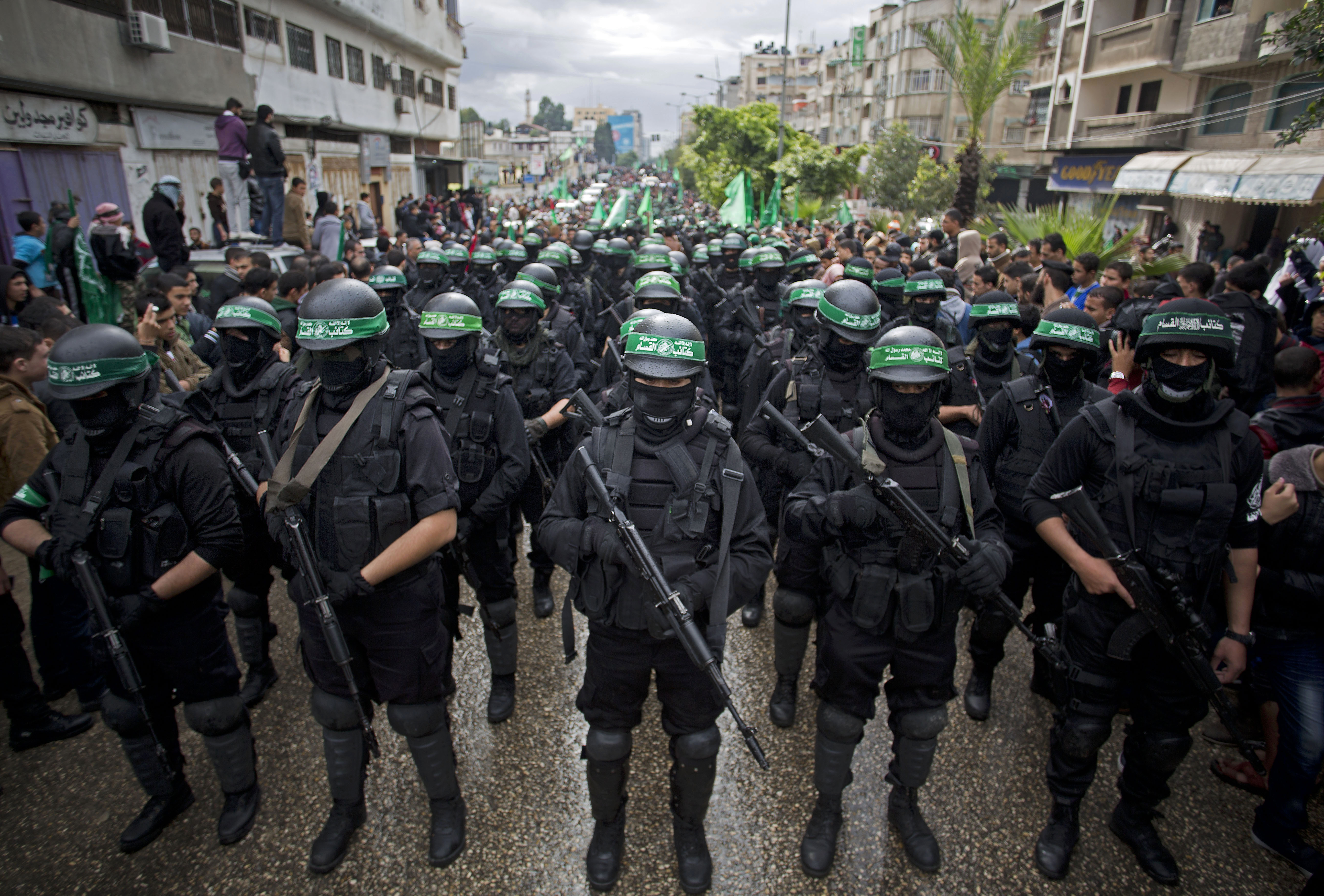 Men in black and olive body armor, their heads covered by helmets bearing the brigades’ green and white logo, their faces masked, march in a deep column down a street, carrying assault rifles.