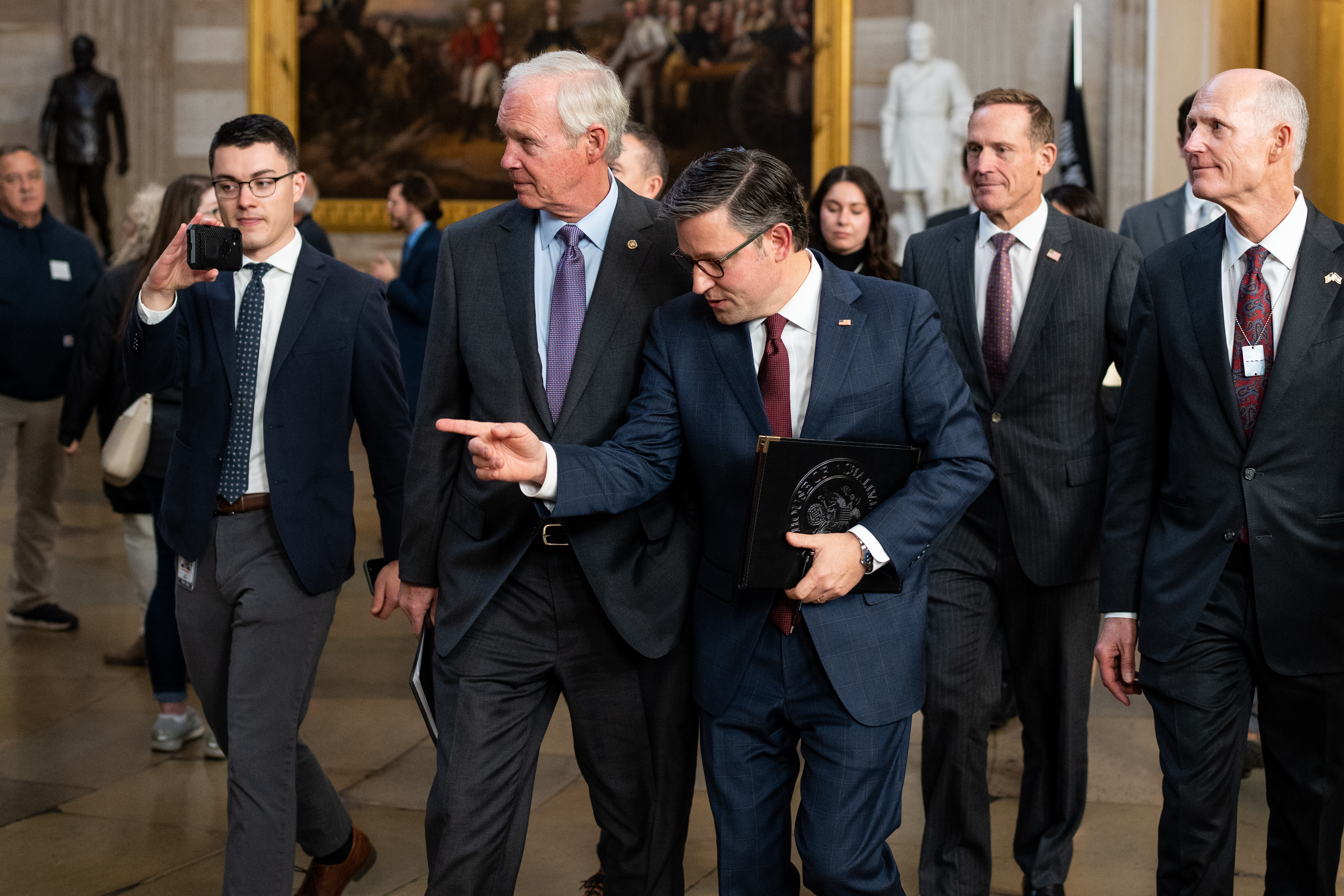 Johnson, clean-shaven in black framed glasses and a dark suit, shoots a finger gun at someone off-camera. He’s flanked by grey and white-haired senators, all in dark blue suits: Sen. Ron Johnson (R-WI), Sen. Ted Budd (R-NC), and Sen. Rick Scott (R-FL).