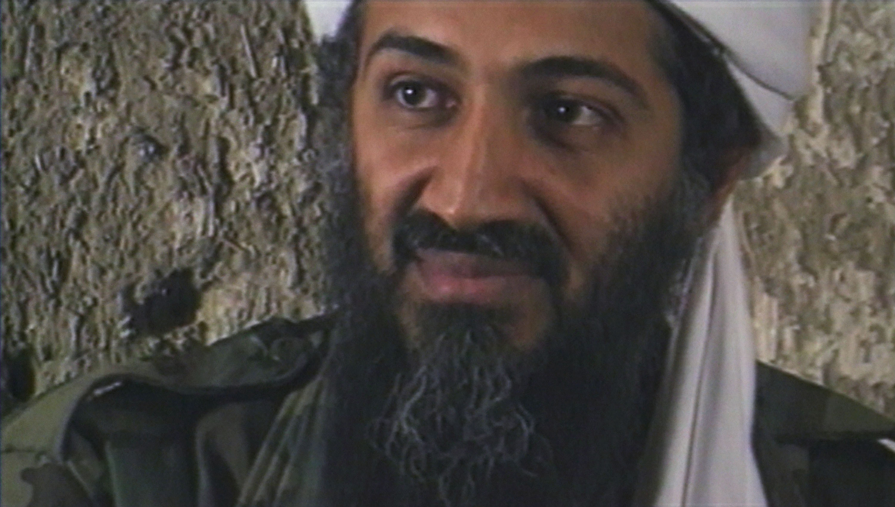 A close-up of bin Laden’s face; it is long, thin. His skin is dark brown, and he sports a thick salt-and-pepper beard as well as a white turban. He appears to be wearing combat fatigues.