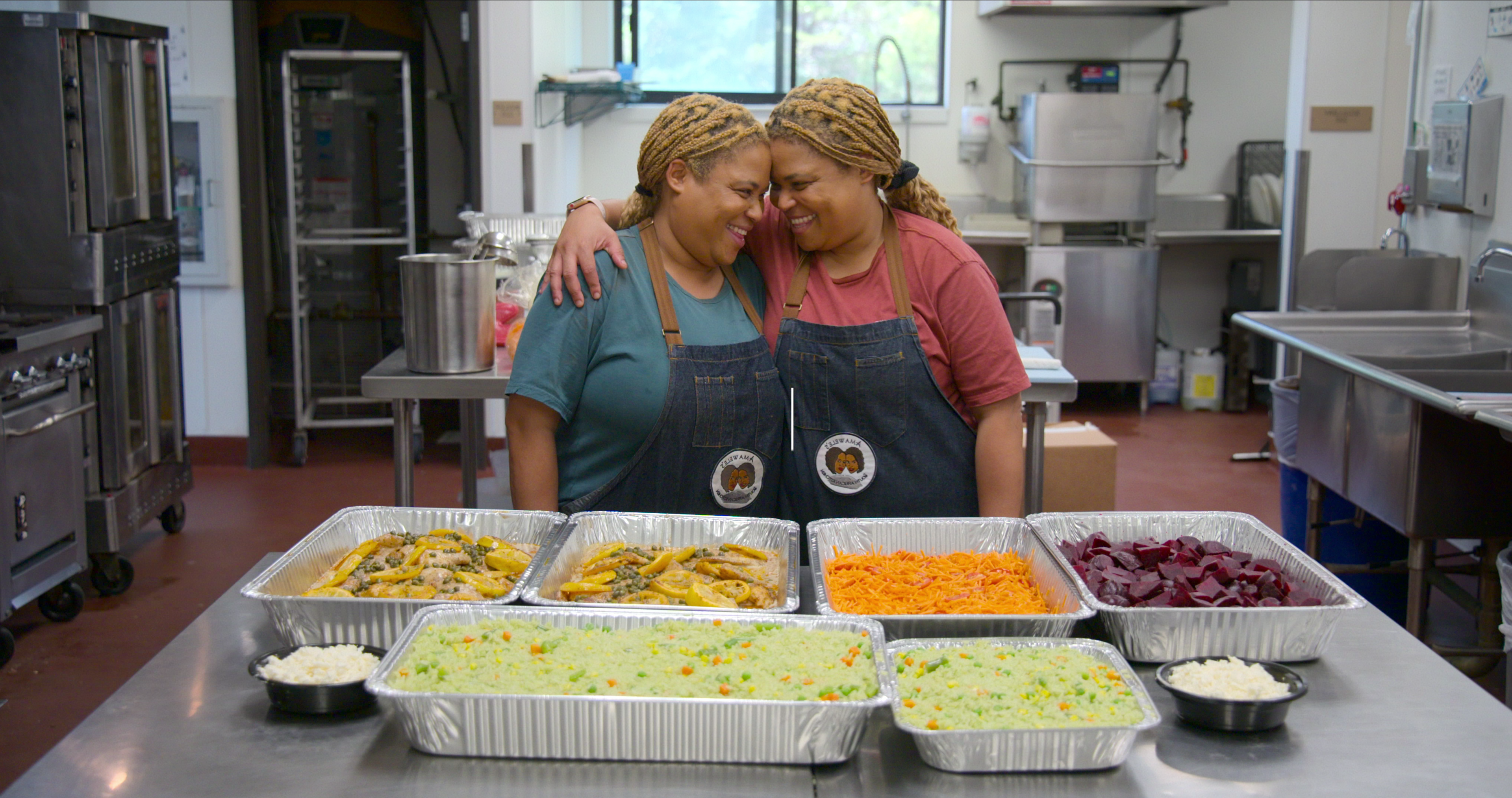 Two women, identical twins, wear aprons and stand in a kitchen with their arms over each other’s shoulders and their heads together. In front of them is a counter with trays of food to be prepped.
