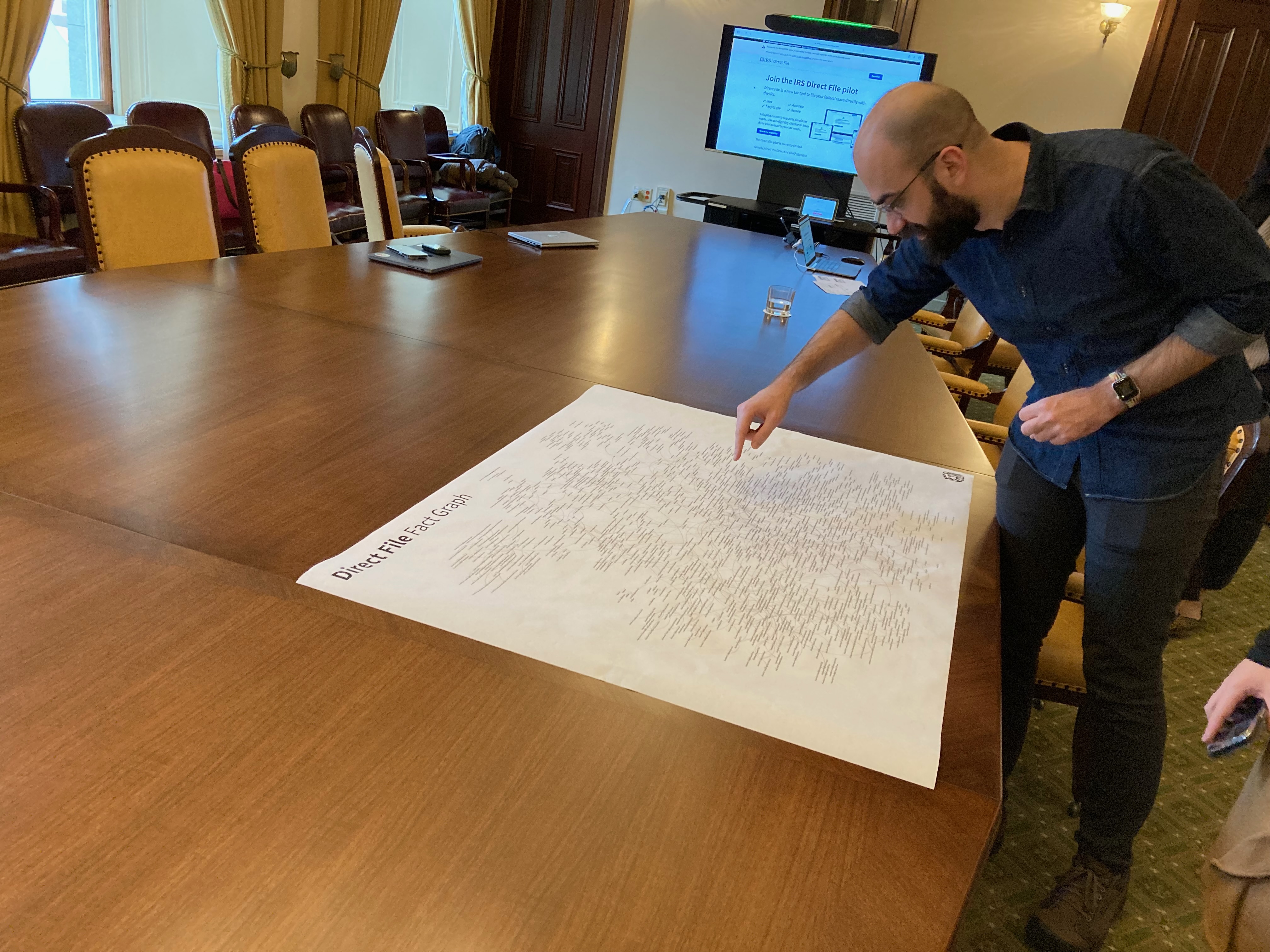 A government software engineer leans over an otherwise empty conference table pointing to a spot on a large board covered in writing.