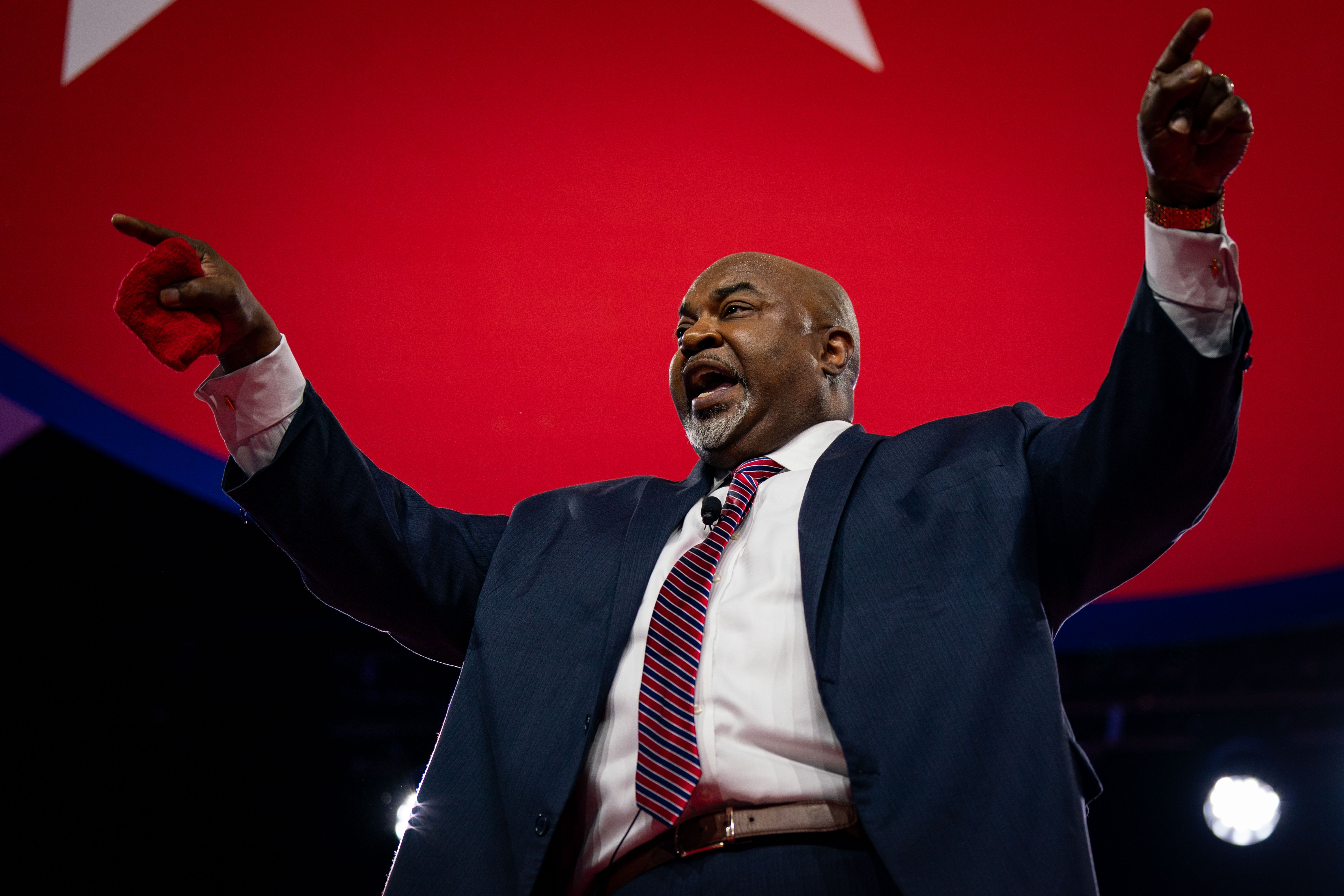 A large Black man in a suit and tie stands onstage with his arms raised while speaking to the audience.