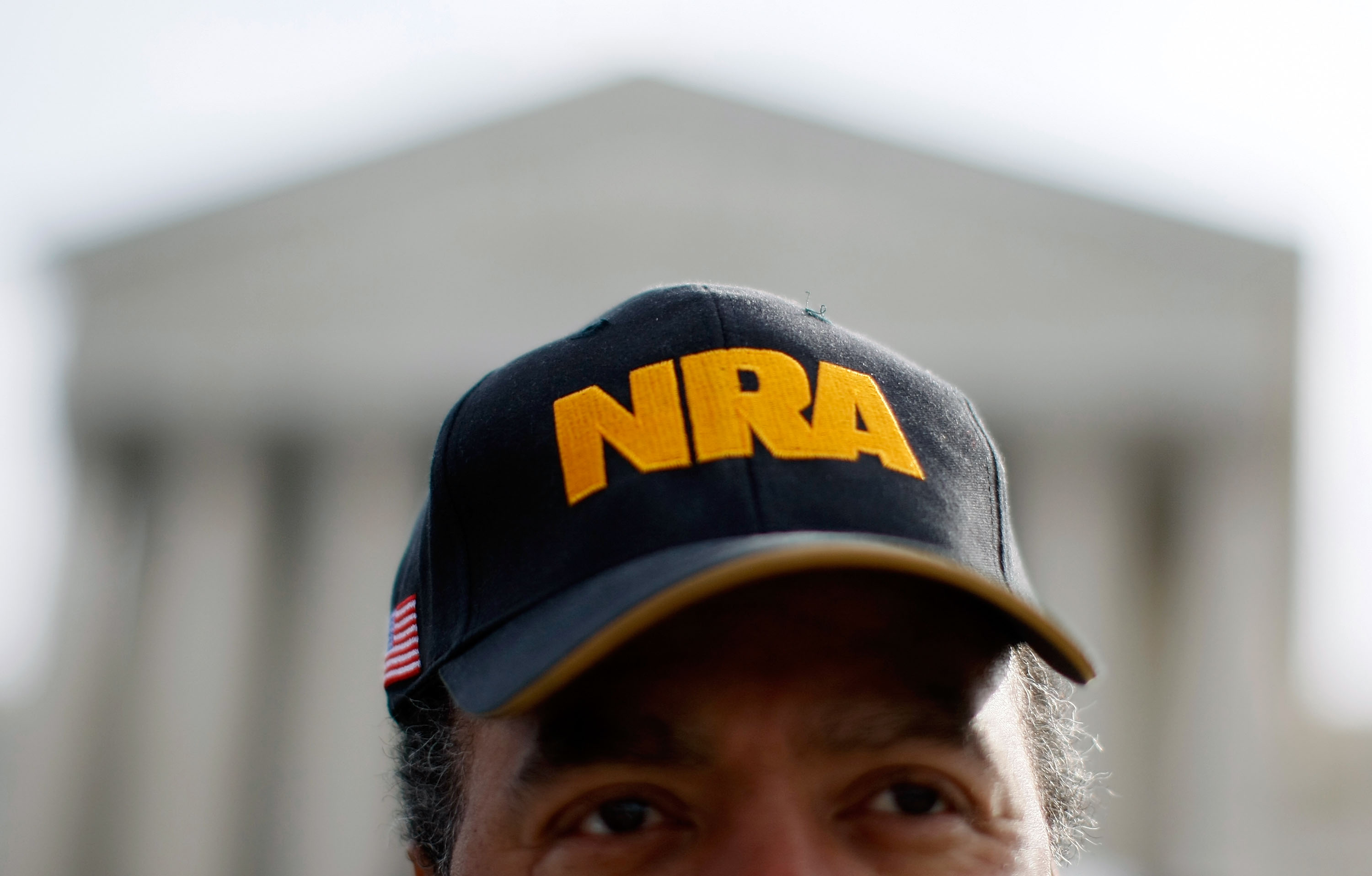 A close-up photo of a man wearing a navy baseball cap embroidered with the letters NRA in yellow. The US Supreme Court building is visible in the background.