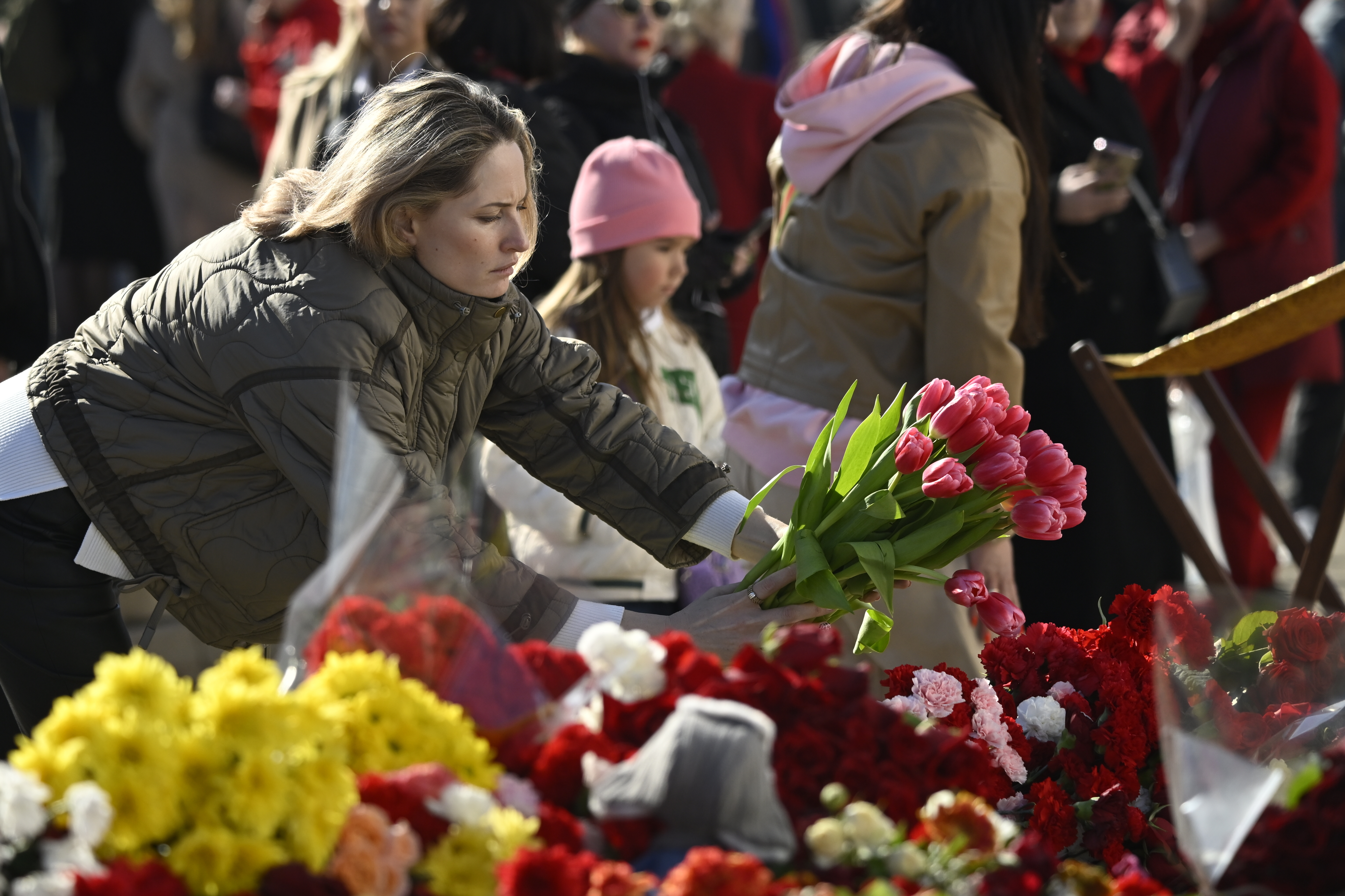 A woman and child are among a crowd of people outside a large concert hall, laying flowers at the base of a pile of flowers, candles, and other memorial tokens. 