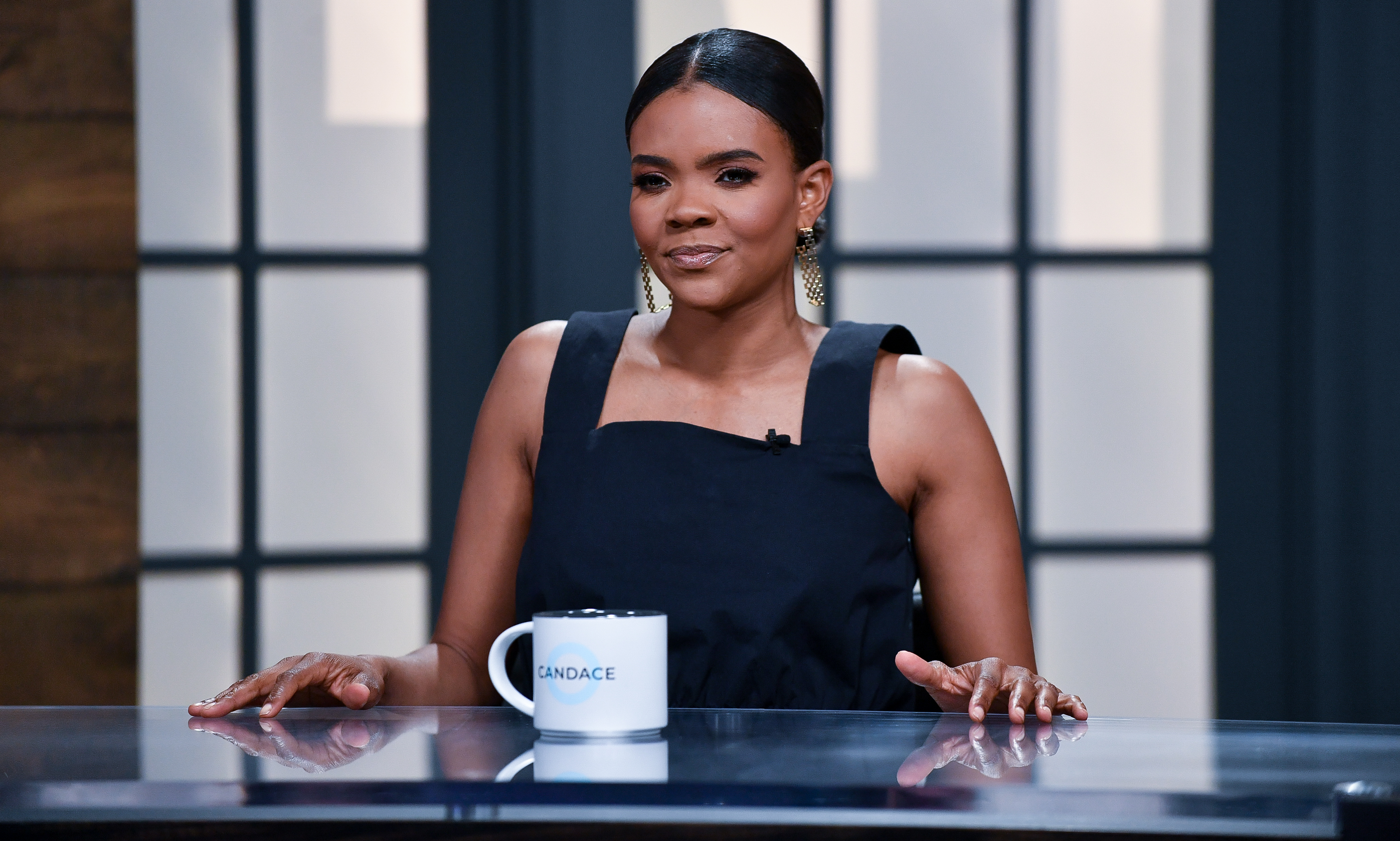 Candace Owens, a Black woman in a black square-necked sleeveless dress with her hair pulled back, sits at a glass desk with a mug reading “Candace” in front of her, and a window behind her.