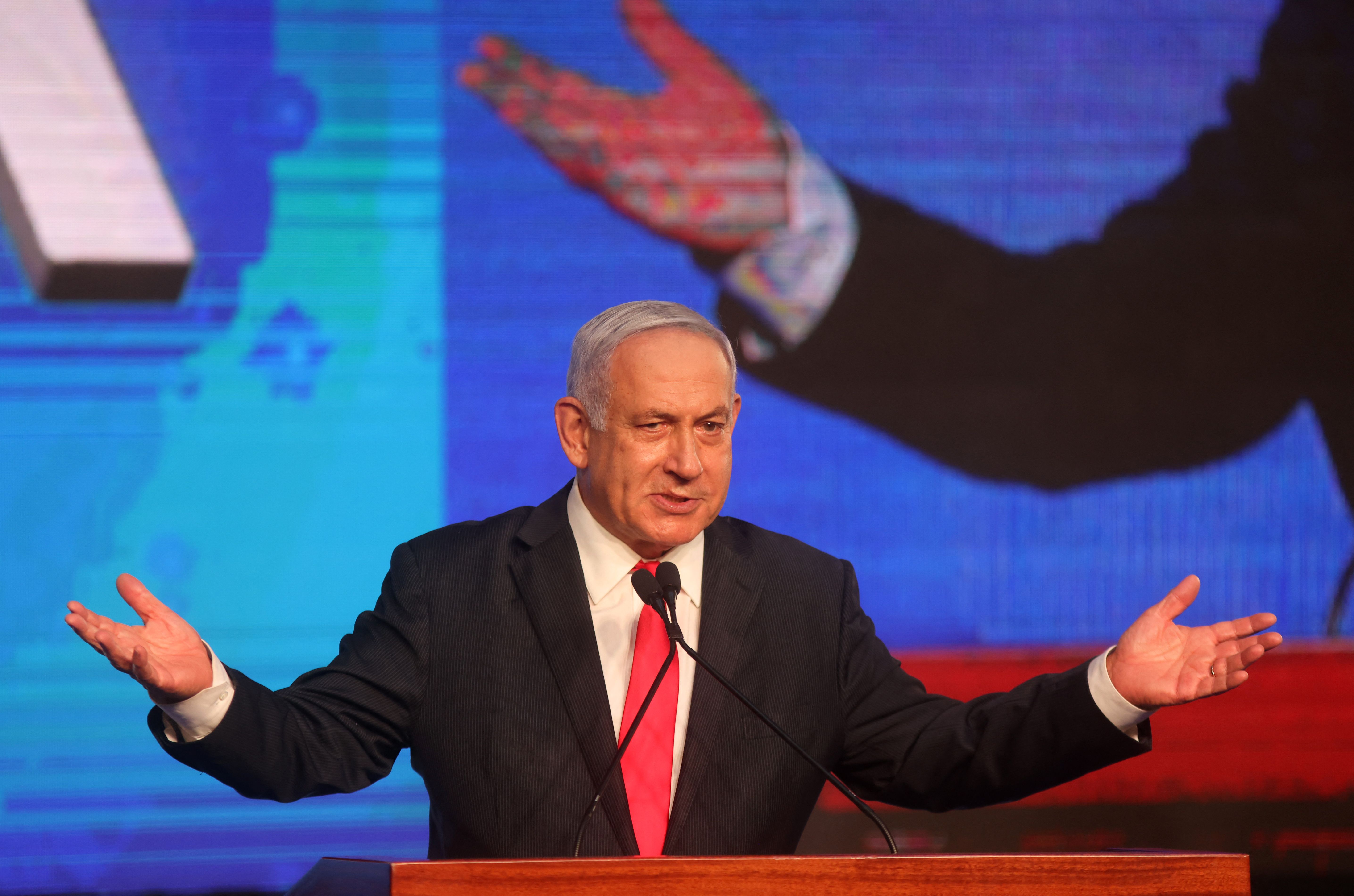 Benjamin Netanyahu, wearing a black suit and red tie, stands in front of a microphone with his arms outstretched. 